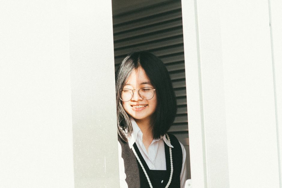 A cheerful woman wearing glasses enjoys the sunlight while standing by a window.