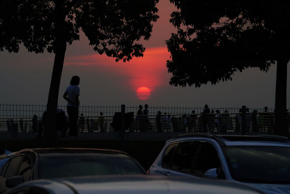 Dramatic sunset view with silhouettes of people relaxing and walking in a park near a parking lot.