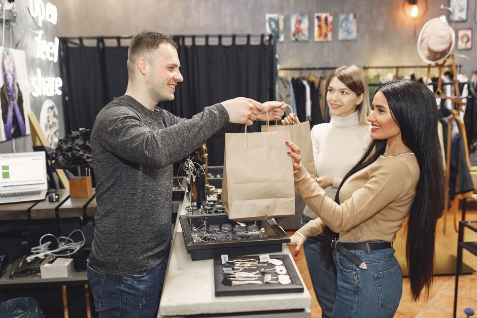 Side view of cheerful male shop assistant standing at counter desk and giving to positive female customers paper bags with purchases in trendy shop
