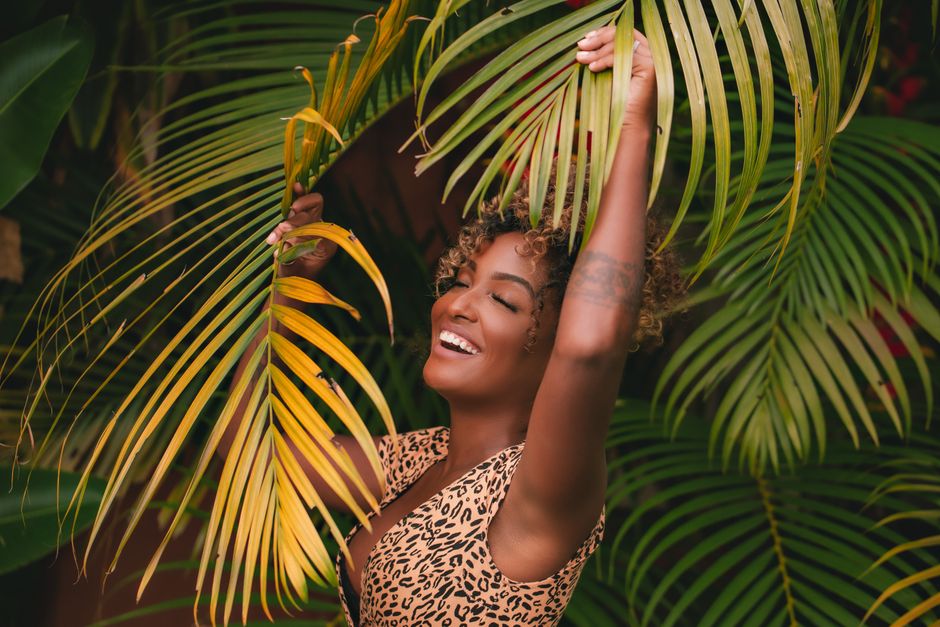 A cheerful woman with curly hair and arms raised, surrounded by vibrant palm leaves, smiling brightly.