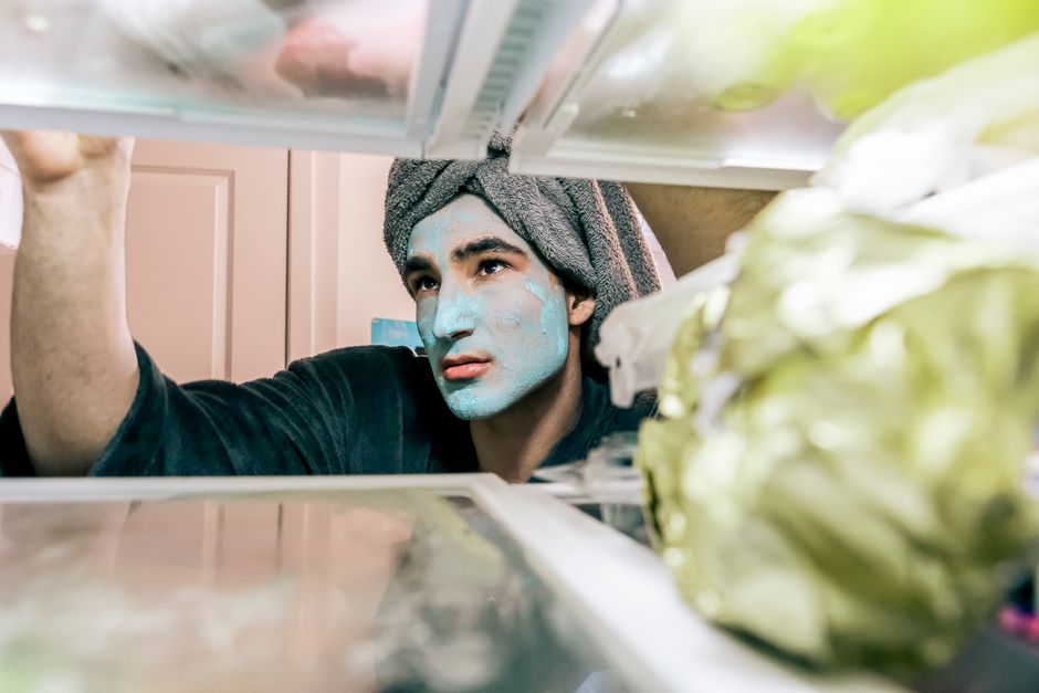 A young man with a blue face mask and towel on his head searches inside a refrigerator.