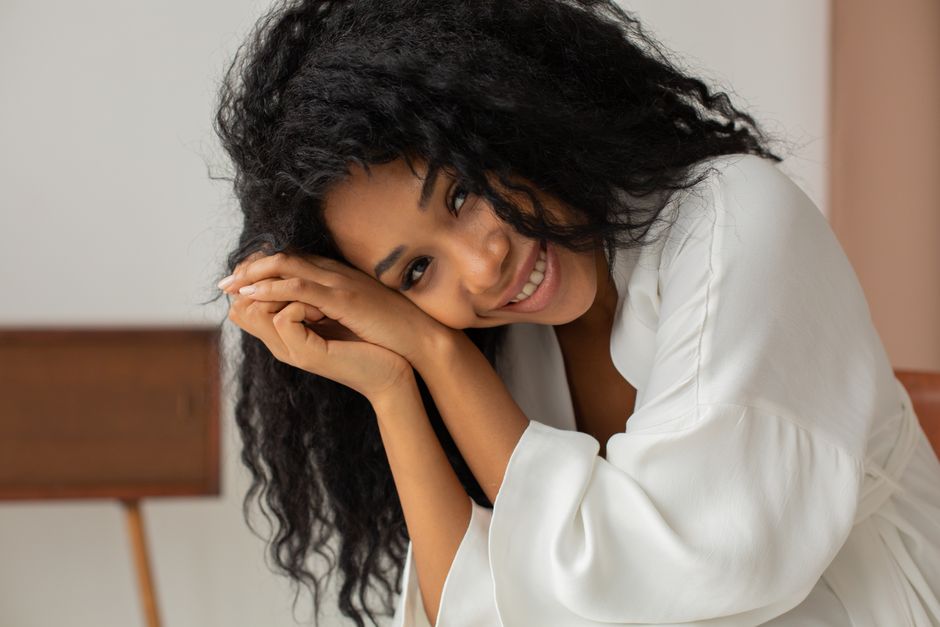 A cheerful woman with curly hair smiling warmly in a white bathrobe, indoors.