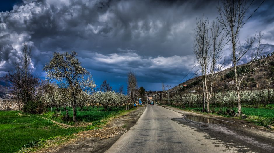 Stunning rural road scene in Kalam, Pakistan with vibrant greenery and dramatic clouds overhead.
