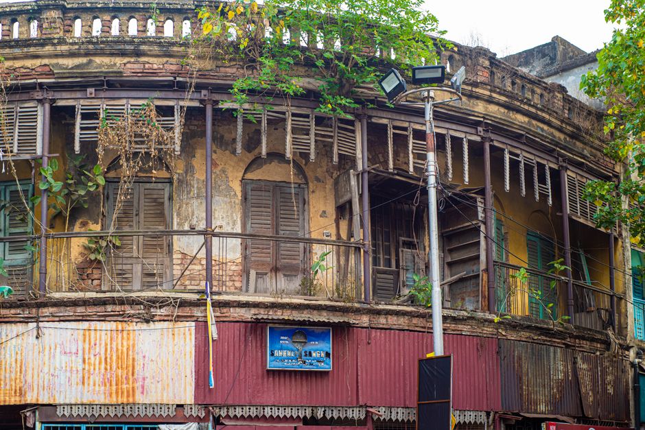 A worn colonial-style building with fading shutters and lush overgrowth in an urban area.