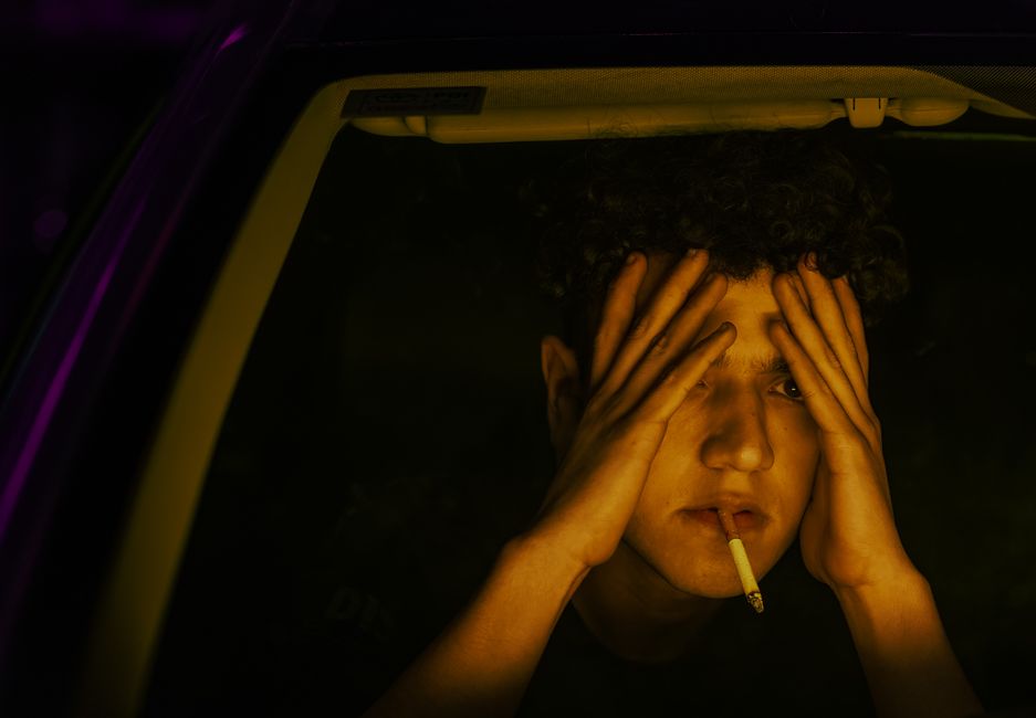 Young man sits with a cigarette, reflecting and pensive, inside a car at night.