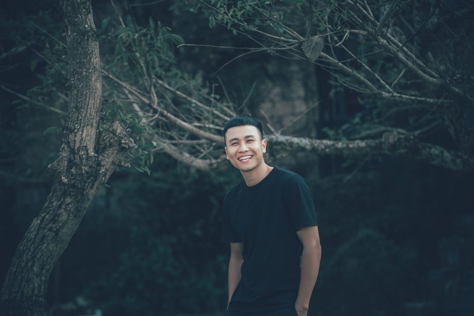 A cheerful man poses outdoors in a natural setting, wearing a black t-shirt and smiling warmly.