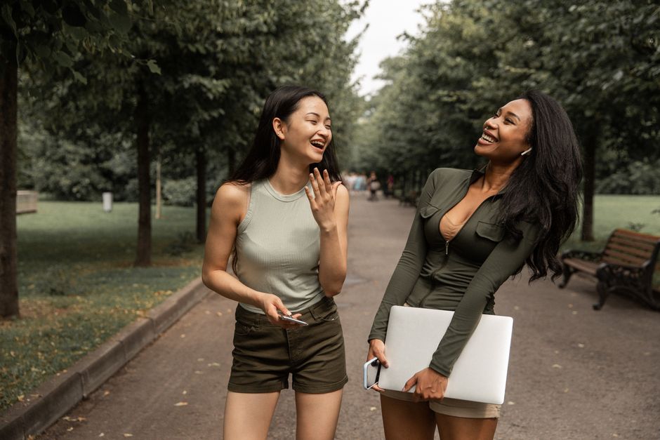 Happy young diverse female friends with smartphone and laptop in hands in casual clothes standing on path amidst lush trees in park and laughing while spending free time together