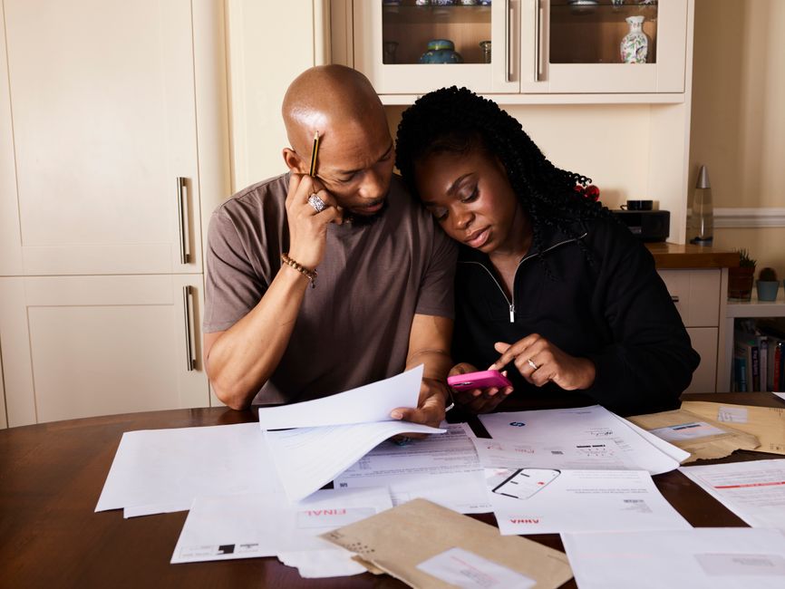 A couple sits at a table managing domestic finances, evaluating documents and using a smartphone.