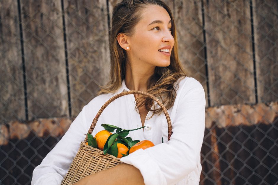 A cheerful woman in a white blouse holds a basket of ripe tangerines outdoors.