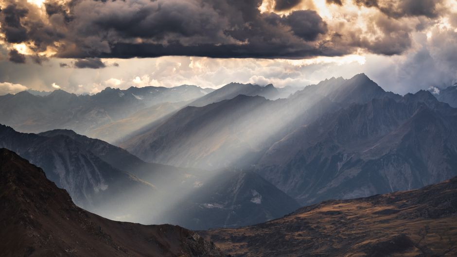 Sun beams through clouds over rugged mountains, creating a dramatic scene.