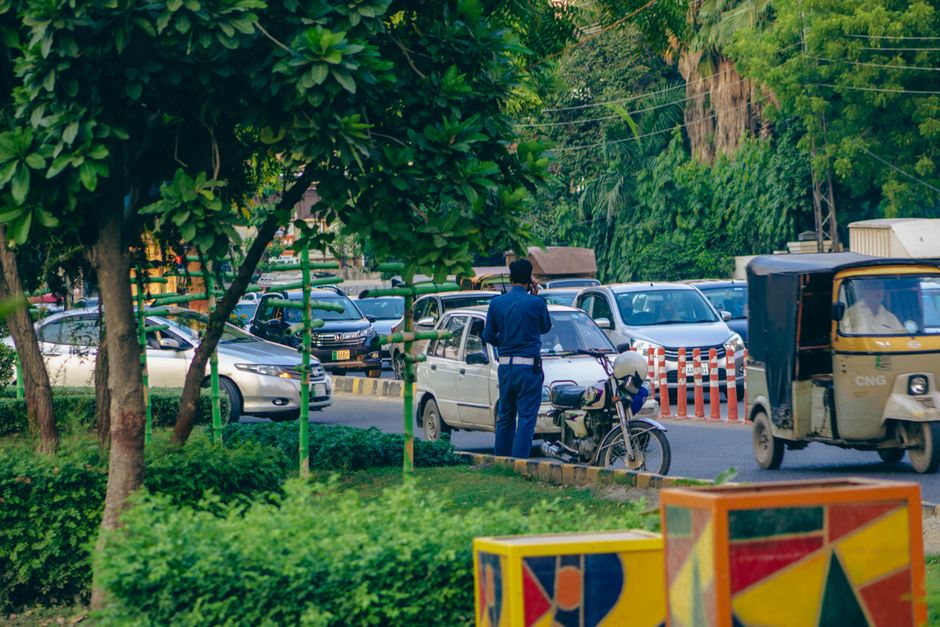 A policeman manages heavy traffic on a bustling street in Lahore, Pakistan, amidst greenery and urban chaos.