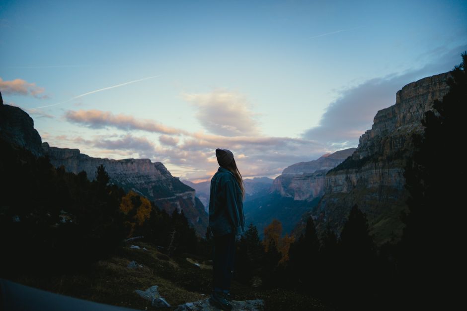A solitary woman stands silhouetted against a stunning mountain landscape at dusk, surrounded by nature.
