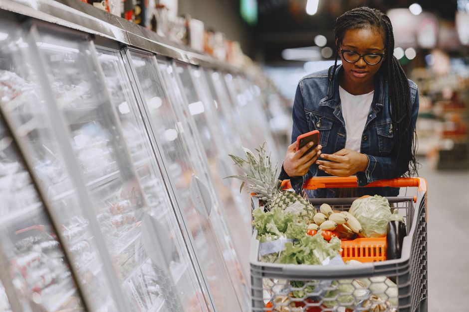 Woman in a grocery store using smartphone while pushing a shopping cart filled with groceries.