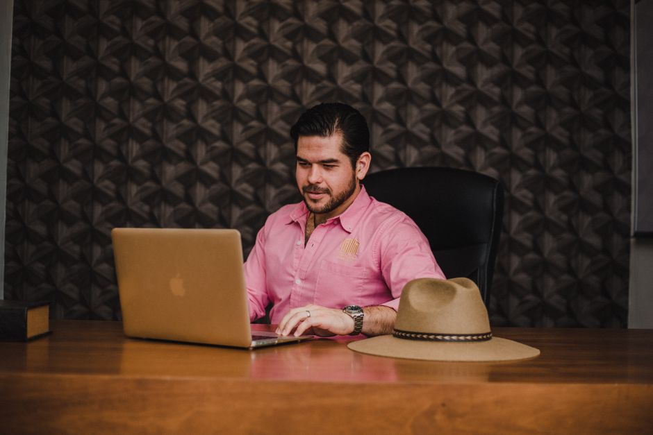 Businessman working on a laptop in an office setting, wearing a pink shirt, with a hat on the desk.