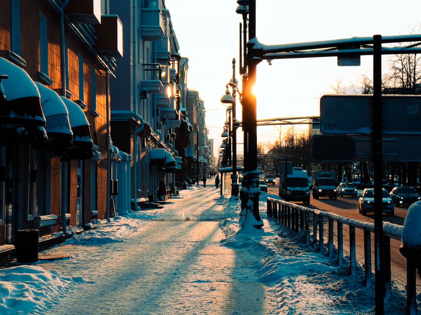 City street at sunset with snow-covered sidewalks and urban traffic, capturing winter ambiance.
