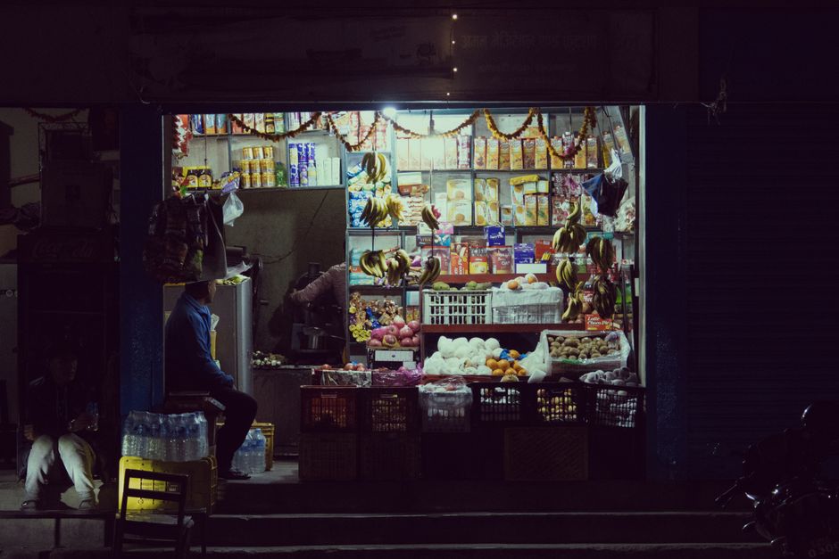 A small street vendor shop at night with illuminated merchandise and groceries.