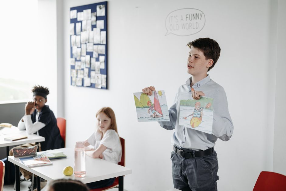 A young student in a classroom presenting colorful drawings to classmates during an art lesson.