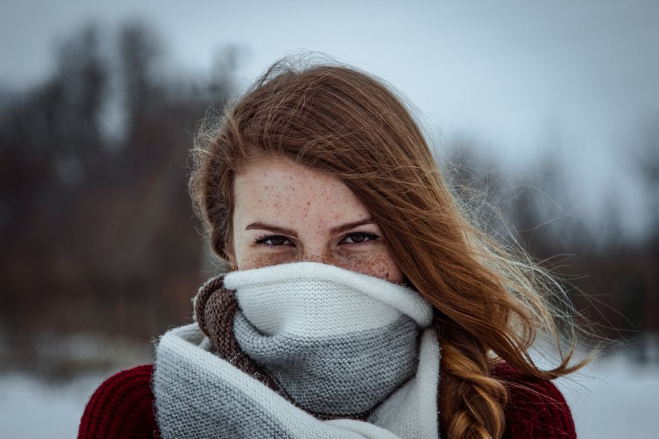Portrait of a young woman with freckles, wrapped in a wool scarf, against a snowy winter background.