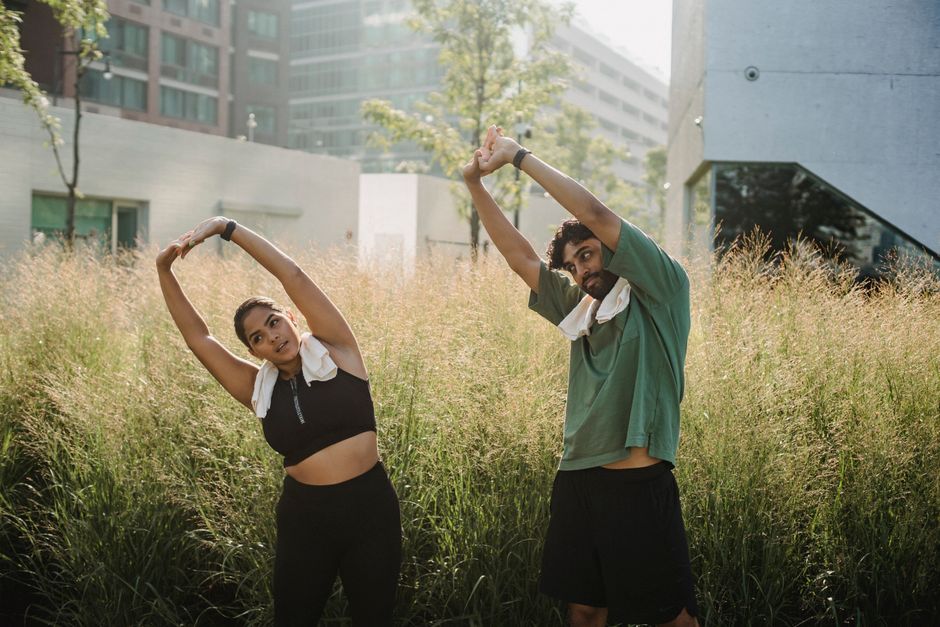 Two young adults perform stretching exercises outdoors in tall grass.