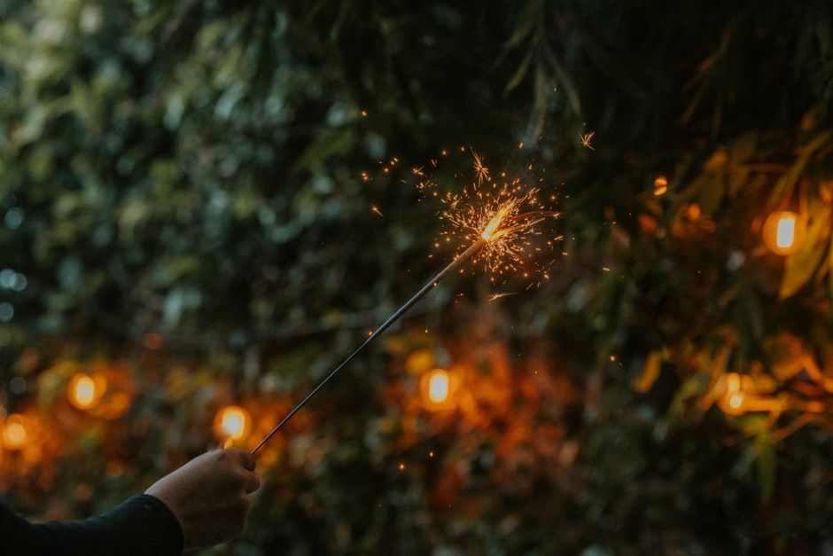 A hand holding a sparker illuminating the night, surrounded by festive outdoor lights.