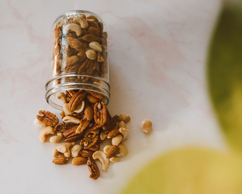 Assorted mixed nuts spill from a glass jar on a light surface, highlighting a variety of textures and colors.