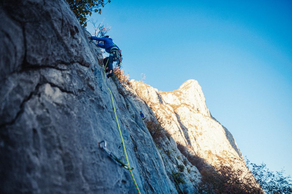 Adventurous rock climber ascending steep mountain face with safety gear in sunlight.