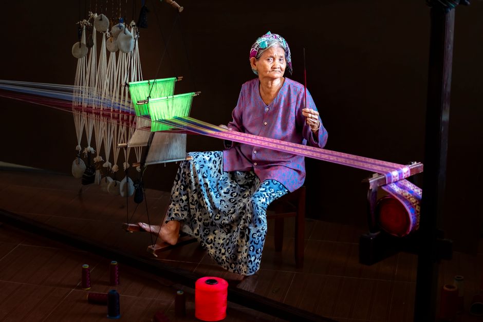 Elderly woman weaving colorful fabric on a traditional loom indoors.
