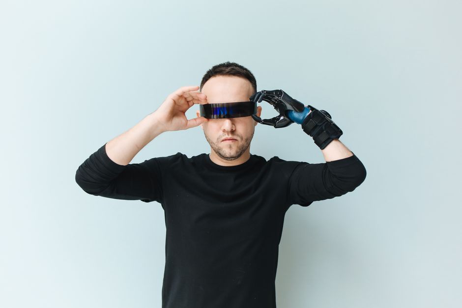 Adult man adjusts futuristic sunglasses with prosthetic arm against neutral background.