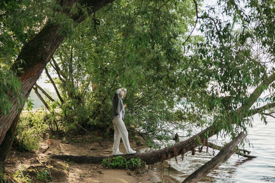 An elderly woman gracefully balances on a fallen tree trunk by a serene lakeside surrounded by lush greenery.