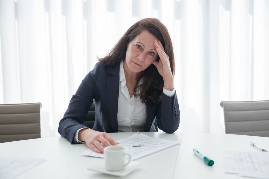 A stressed businesswoman in corporate attire sits at a desk with papers, looking overwhelmed.