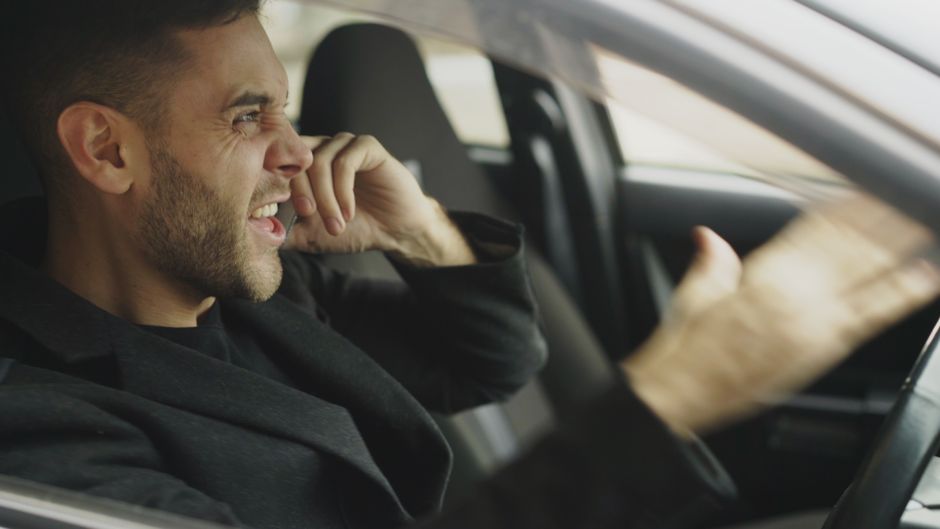 An adult man expressing frustration while on the phone inside a car.